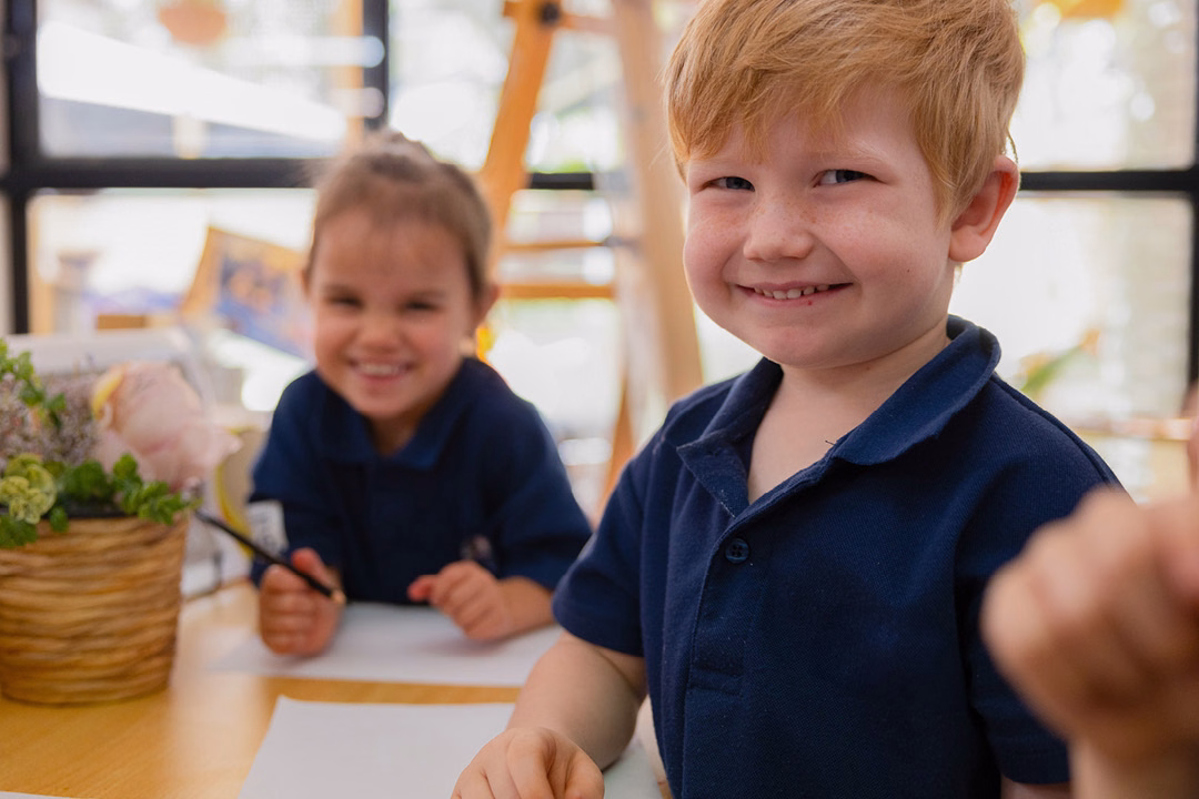 Two children in navy school polo shirts smiling at a table with paper and pencils